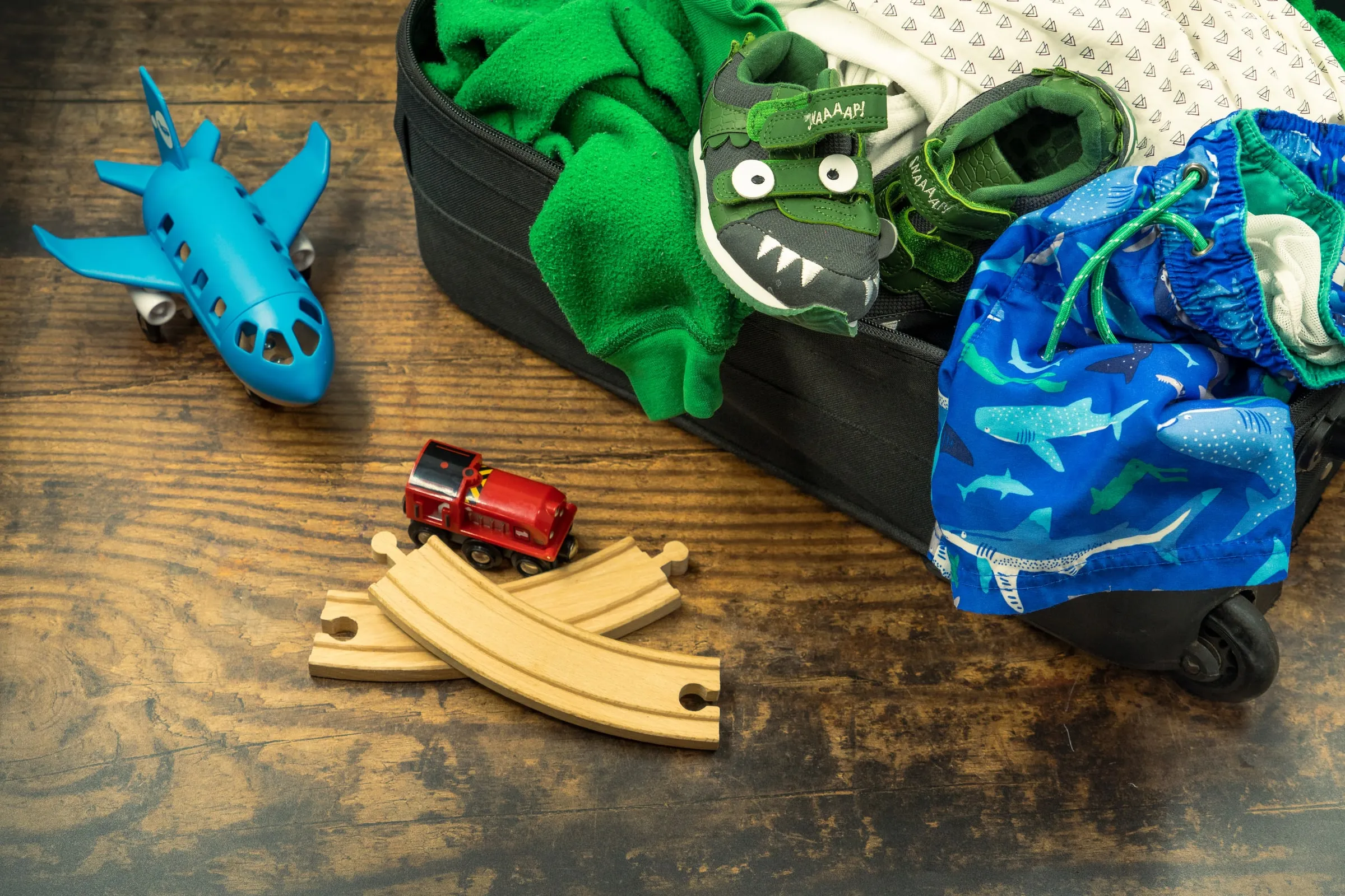 Halb gepackter Kinderkoffer auf Holzdielen, daneben ein Spielzeugflugzeug, eine Holzeisenbahn und Badeshorts