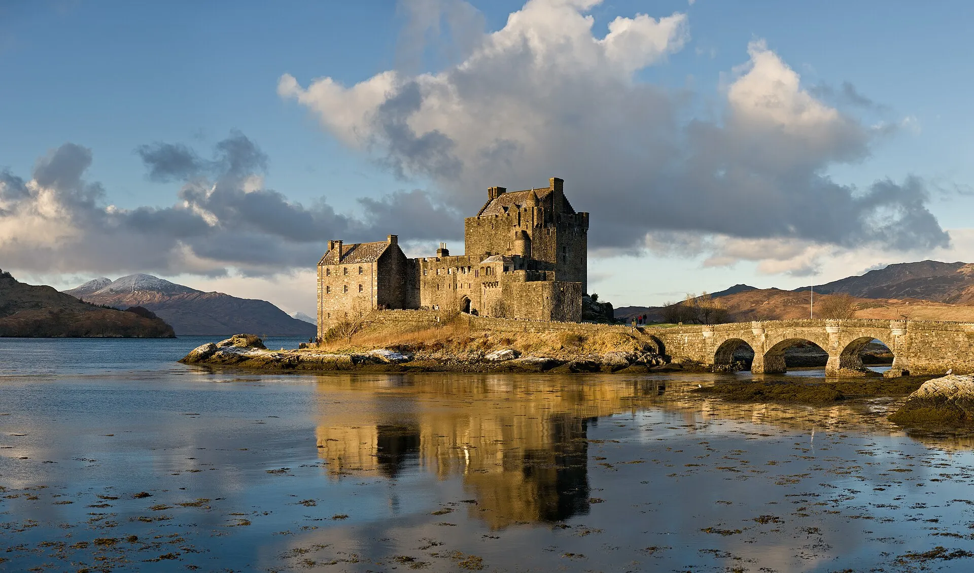 Eilean Donan Castle mit Bogenbrücke im Morgenlicht, gespiegelt im Wasser, dahinter Berge mit Schnee