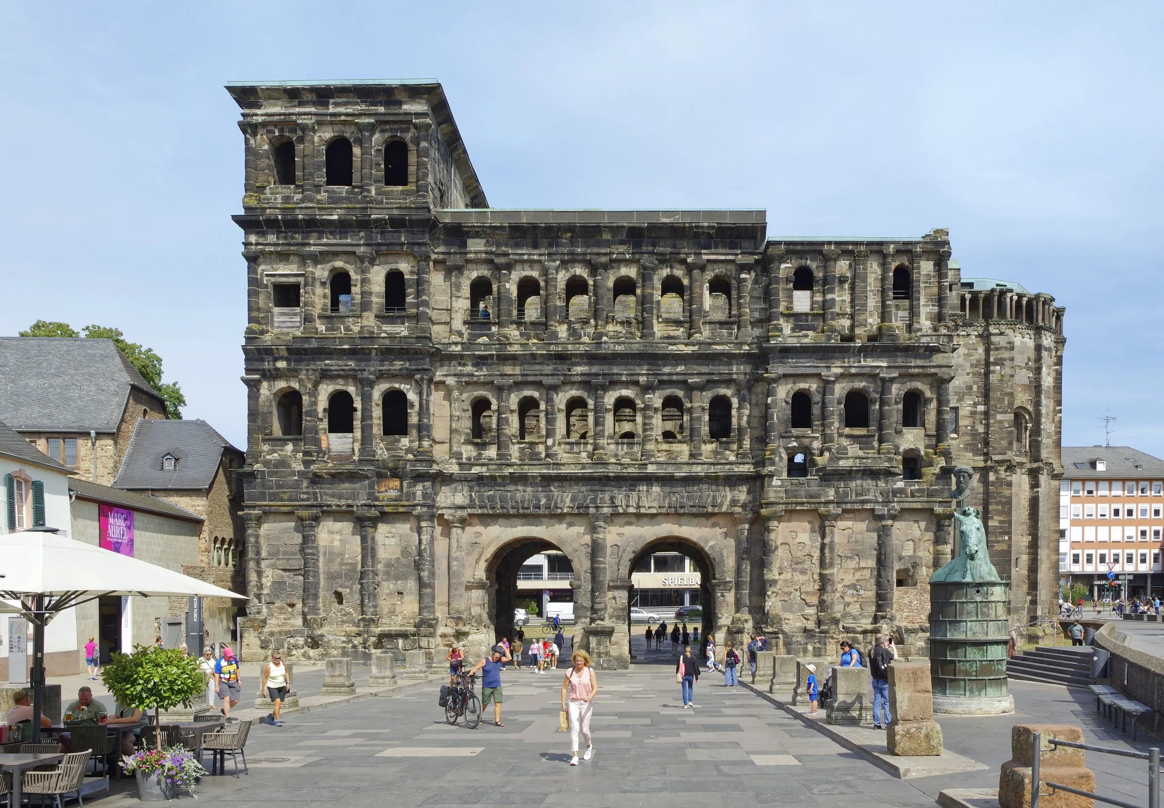 Dunkle Sandsteinfassade der Porta Nigra in Trier mit Torbögen und Fensterreihen, davor ein Platz mit Fußgängern und Café