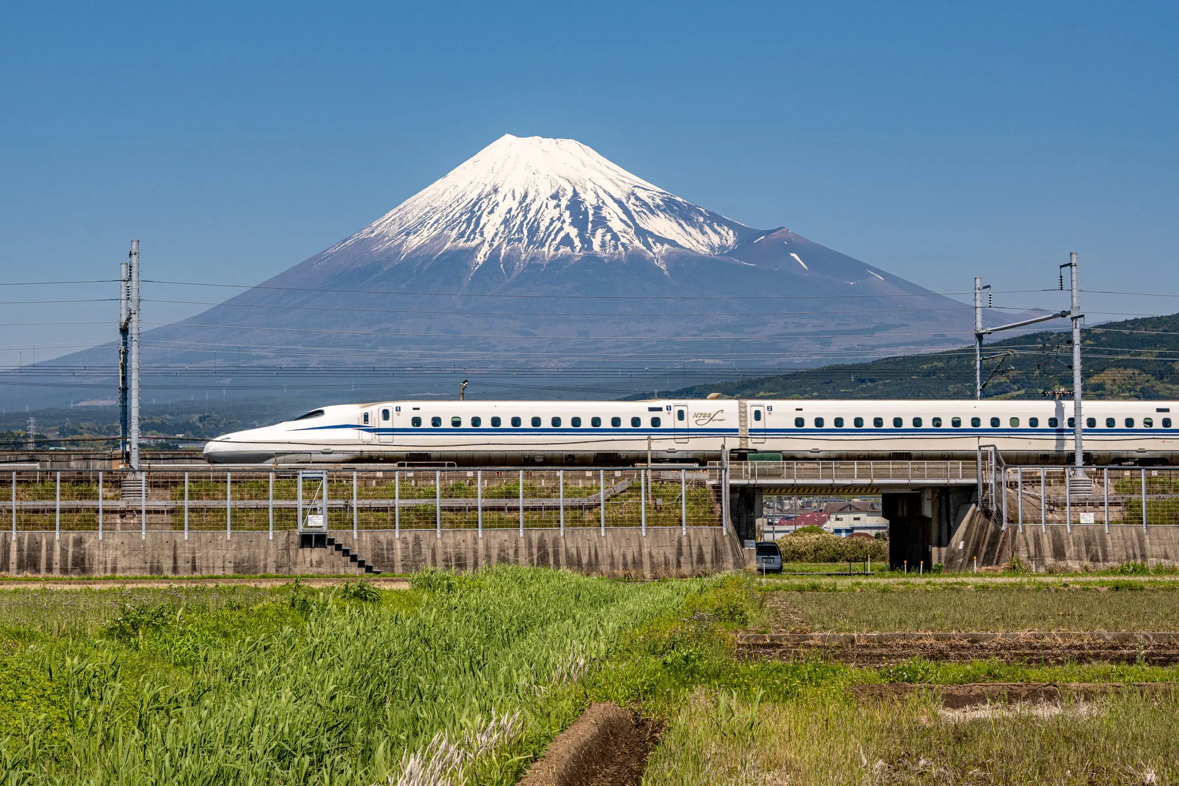 Weißer Shinkansen auf einem Damm zwischen grünen Feldern, dahinter der schneebedeckte Kegel des Fuji