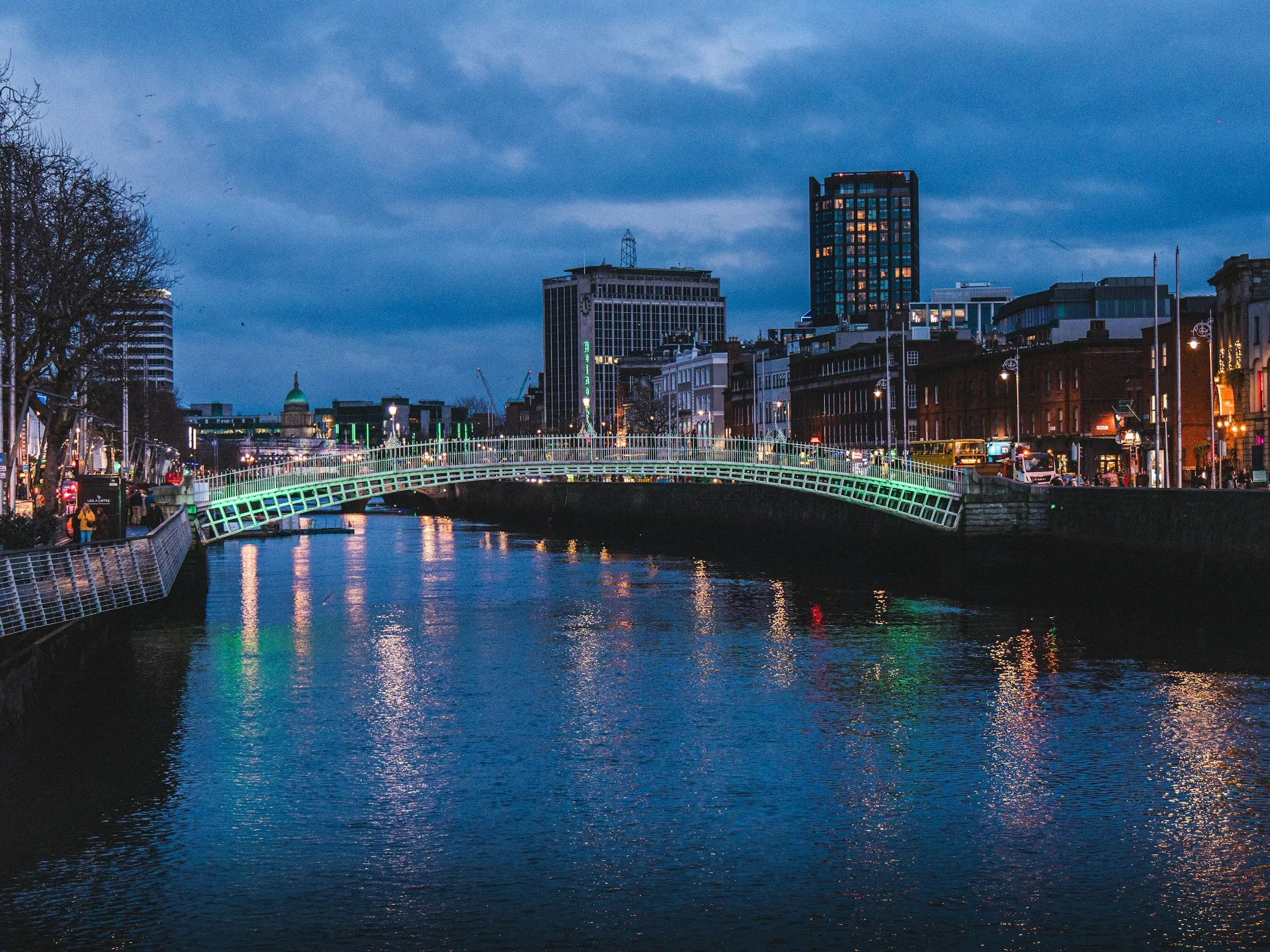 Grün beleuchtete Ha'penny Bridge über der Liffey in der Abenddämmerung, dahinter Bürotürme und eine grüne Kuppel