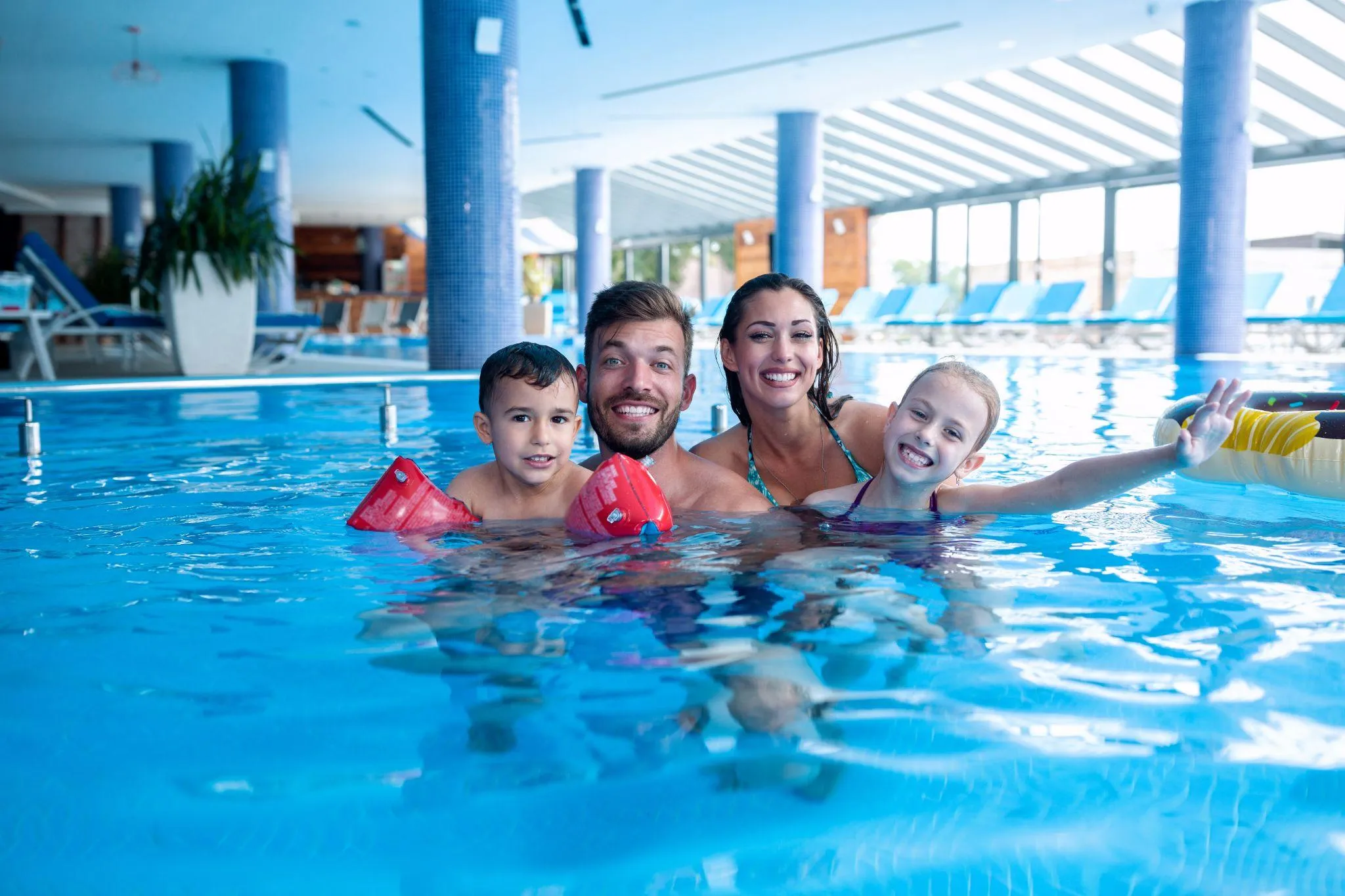 Familie mit zwei Kindern lacht im Becken eines Hallenbads, der Junge trägt rote Schwimmflügel