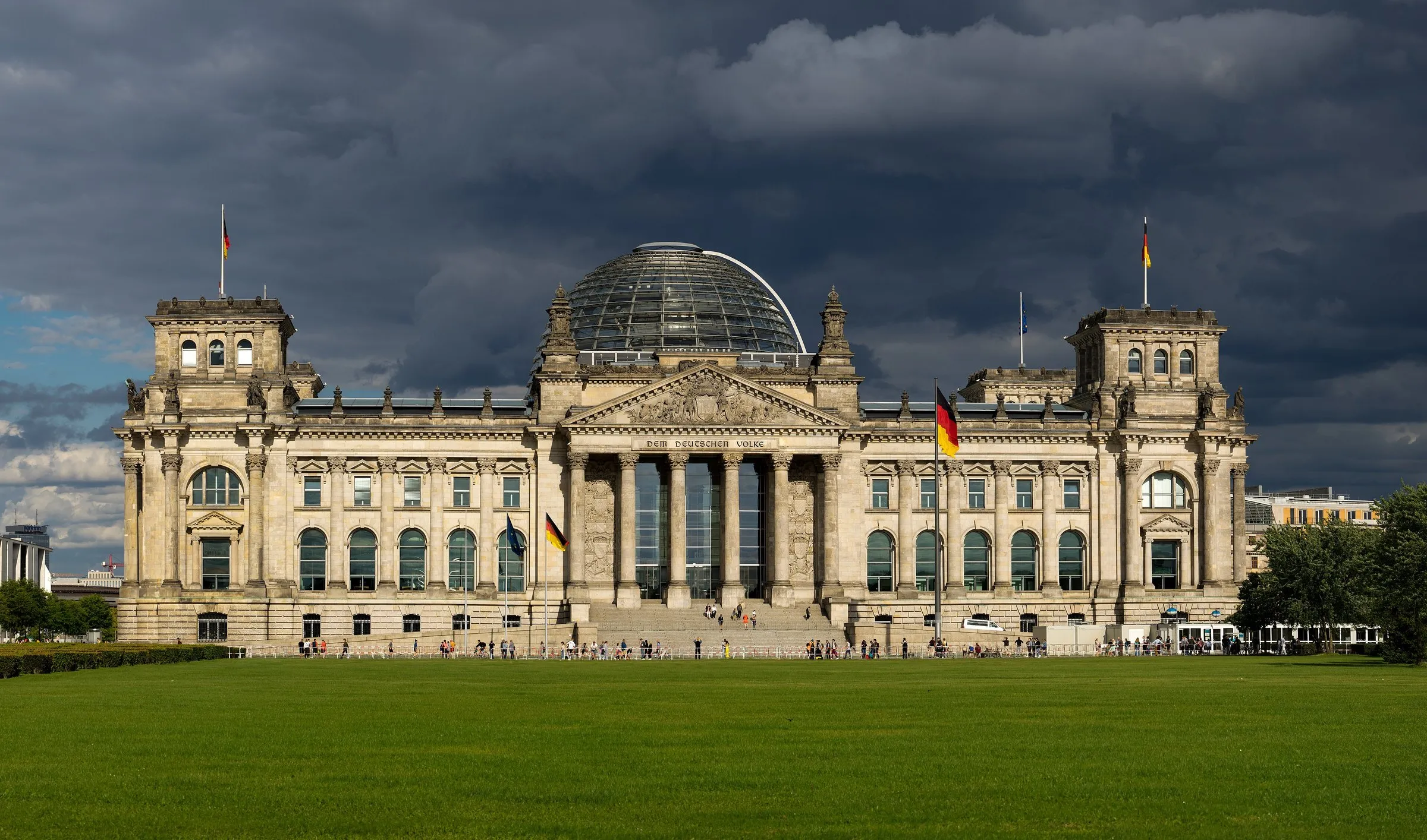 Westfassade des Reichstagsgebäudes mit Glaskuppel über der Wiese des Platzes der Republik unter dunklem Wolkenhimmel