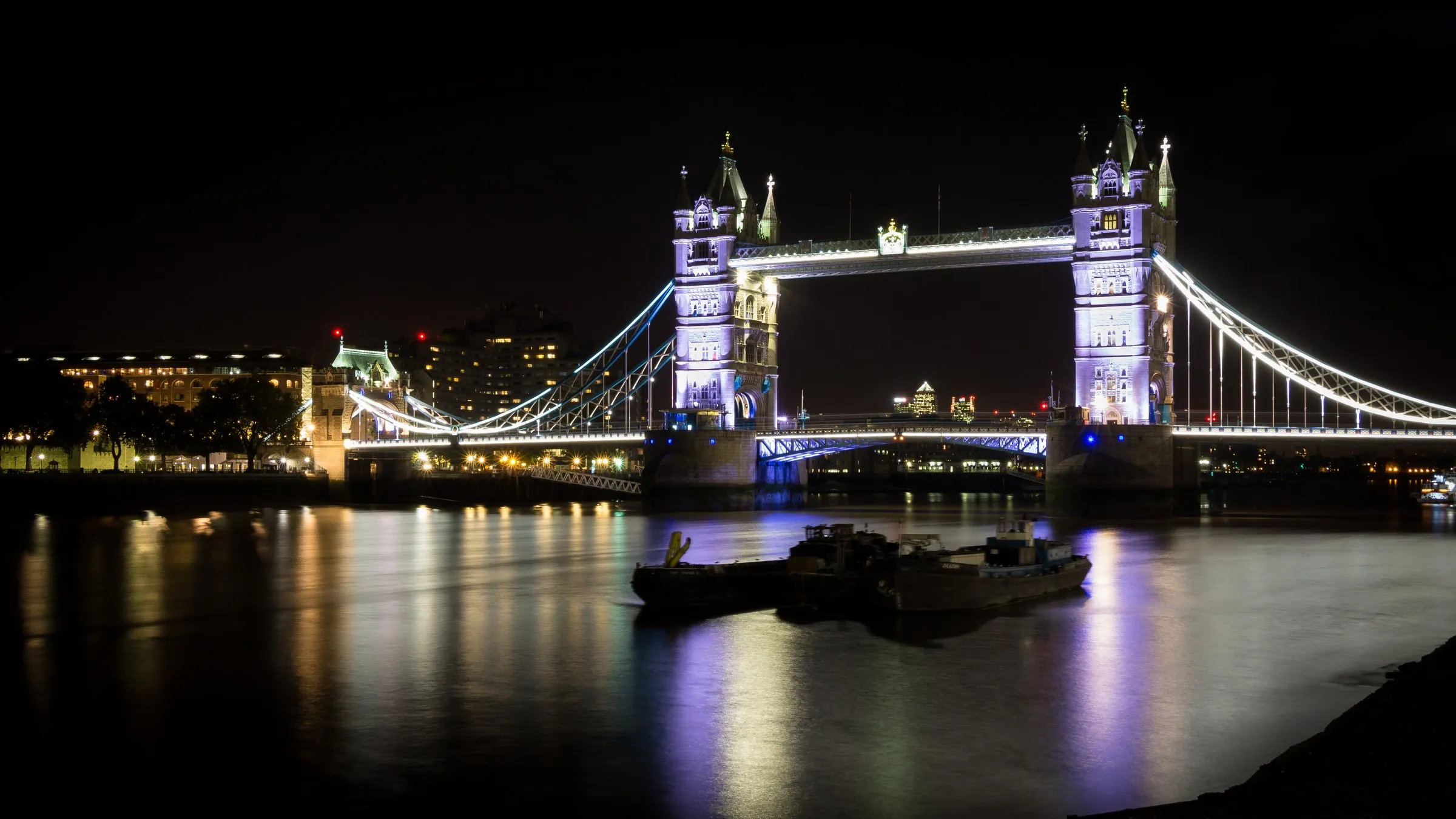 Beleuchtete Tower Bridge über der Themse bei Nacht, davor ein Lastkahn, die Lichter spiegeln sich im Wasser