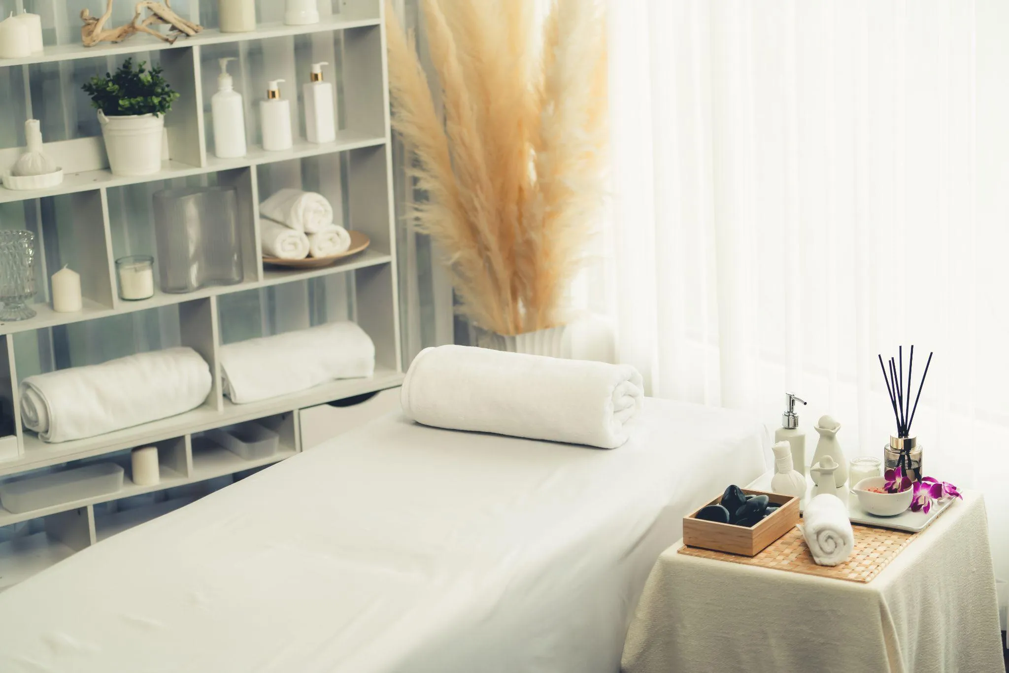 Liege mit weißem Handtuch in einem hellen Behandlungsraum, daneben Massagesteine, Orchidee und Duftstäbchen