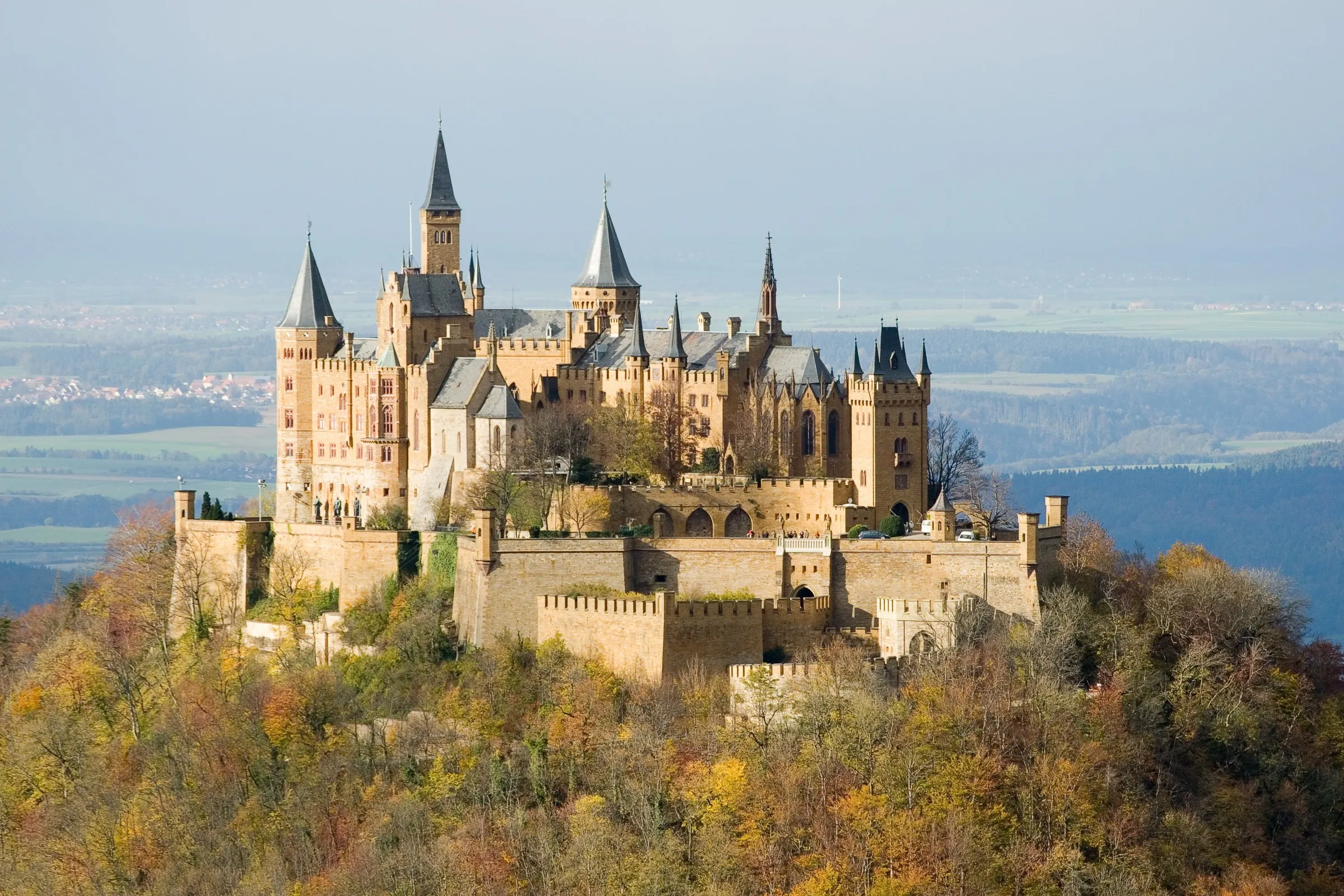 Burg Hohenzollern mit Türmen und Mauern auf dem bewaldeten Berg, das Laub herbstlich gefärbt, dahinter dunstiges Flachland