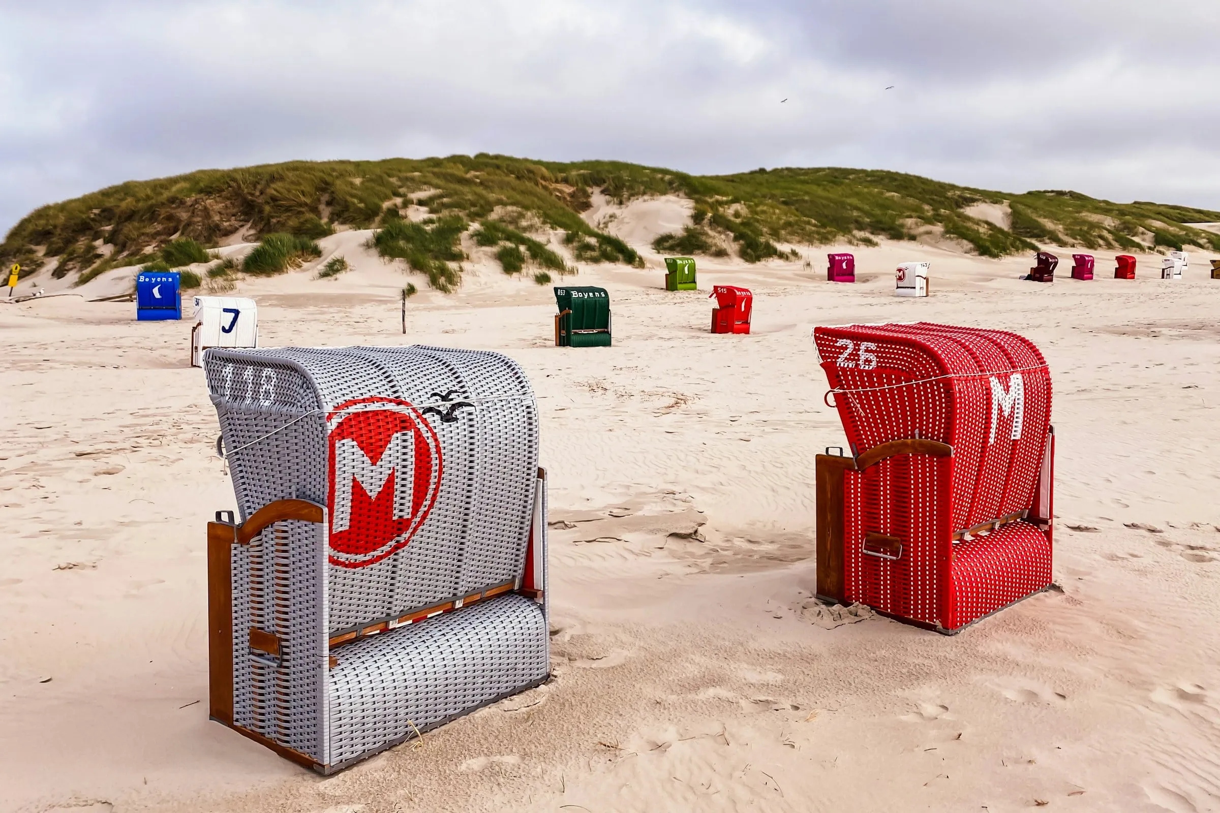 Bunte Strandkörbe auf dem hellen Sandstrand bei Norddorf auf Amrum, dahinter grasbewachsene Dünen unter grauem Himmel