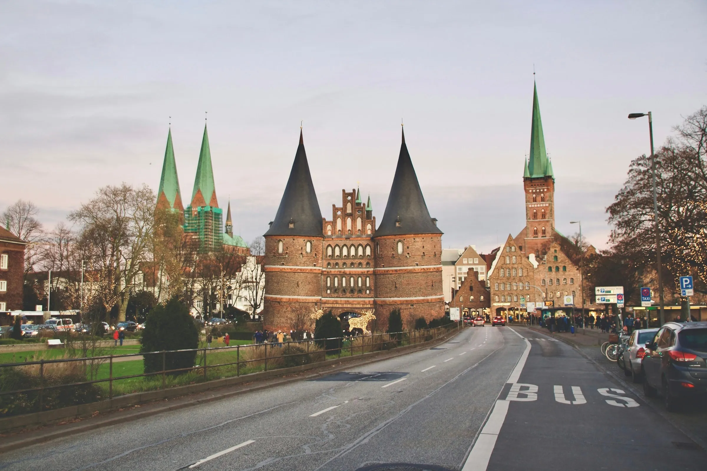 Das Holstentor in Lübeck mit zwei spitzen Türmen, dahinter grüne Kirchturmspitzen der Altstadt, vorn eine Straße