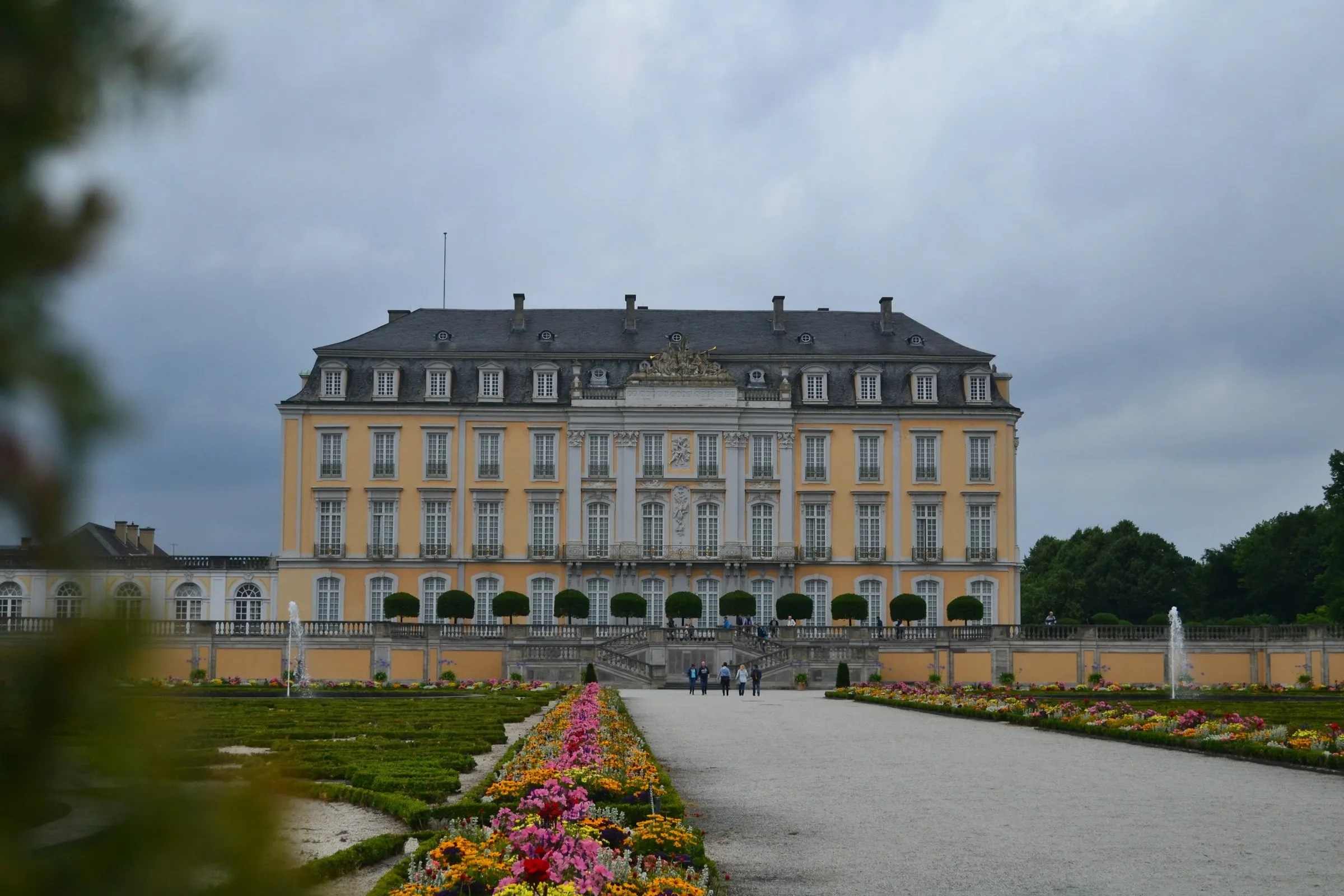 Schloss Augustusburg in Brühl mit gelber Fassade, davor der Barockgarten mit Blumenbeeten und breitem Kiesweg