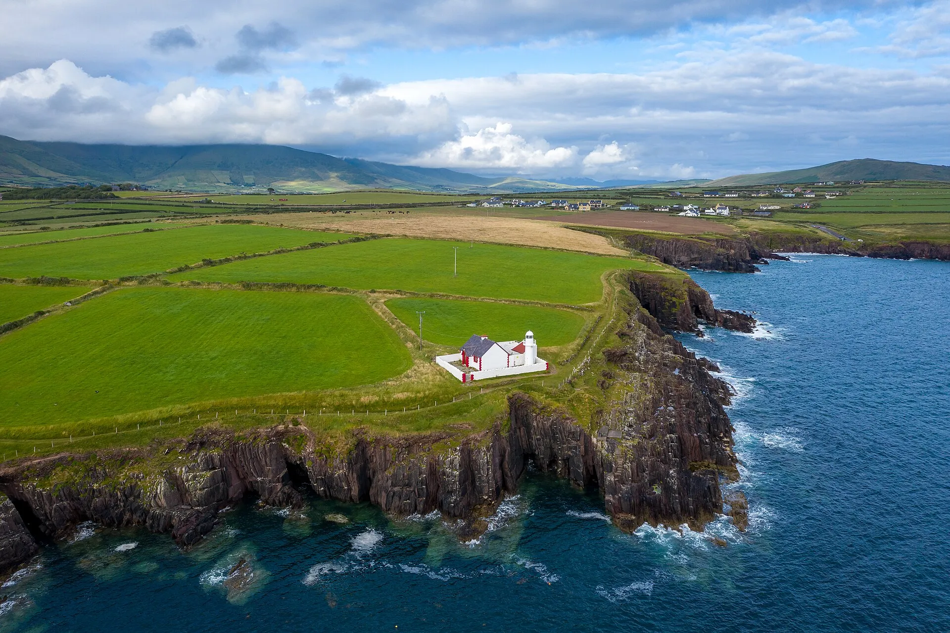 Weißer Leuchtturm auf einer grünen Klippe an der Einfahrt zur Dingle Bay, dahinter Felder und Berge unter Wolken