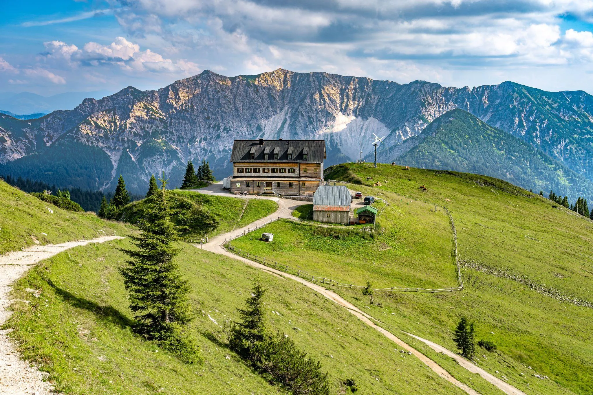 Berghütte aus Holz auf einer Almwiese mit Wanderwegen, dahinter eine schroffe Felswand der Voralpen
